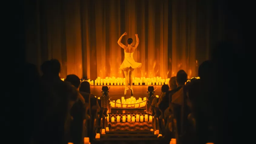 A ballet dancer in a white dress performs on a dimly lit stage, illuminated by the warm glow of numerous candles. The audience watches from candle-lit aisles, creating an intimate and atmospheric setting.
