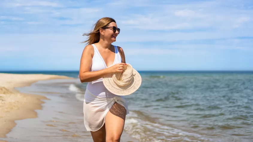 Woman in bathing suit with hat walking along beach in front of at Sanibel Arms West Condos