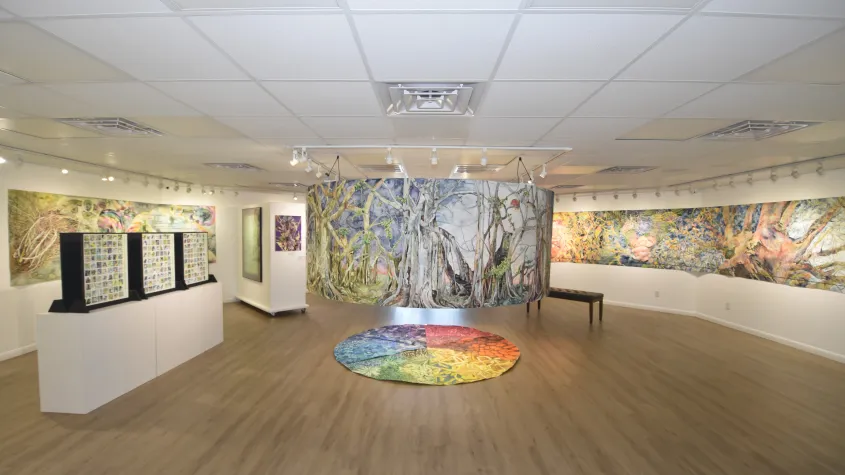 Large gallery installation featuring colorful, nature-inspired artwork covering the surrounding walls and a curved freestanding display in the center. A circular multicolored artwork rests on the floor beneath the installation, with additional artwork displayed on white pedestals nearby.