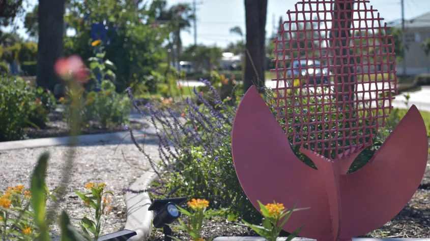 Pink metal sculpture with a grid-like upper section and curved leaf-shaped forms displayed among flowering plants in a sunny landscaped garden.