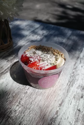 Clear plastic bowl of acai topped with sliced strawberries, banana, coconut flakes, seeds, and granola on a sunlit wooden table.