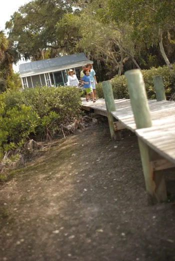 Family walking along a wooden boardwalk through lush coastal greenery toward a small cottage-style home.