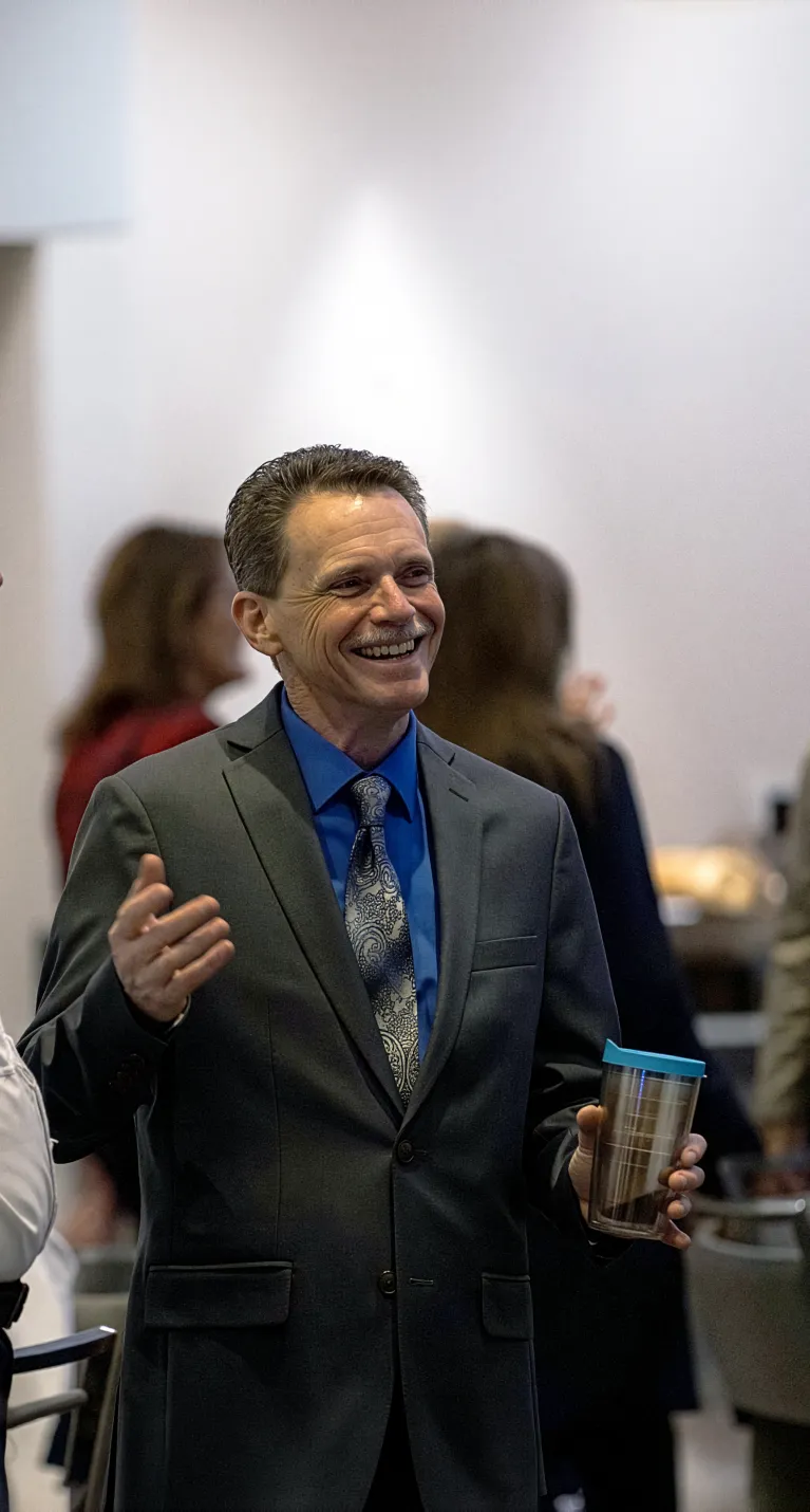 Two men in suits smile and talk during a networking moment at an indoor professional event, with other attendees conversing in the background.