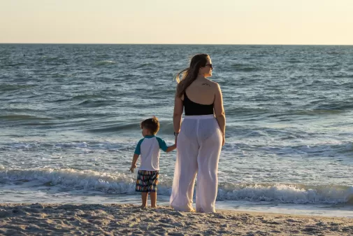 Woman and child holding hands at the water’s edge on a beach at sunset.