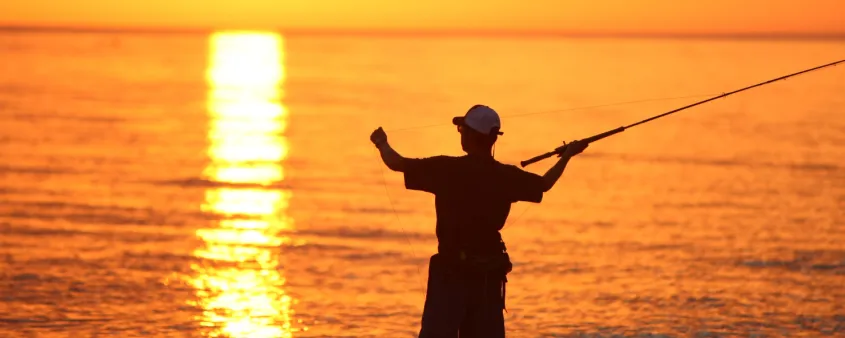 A man casts his fishing line from the shore at sunset