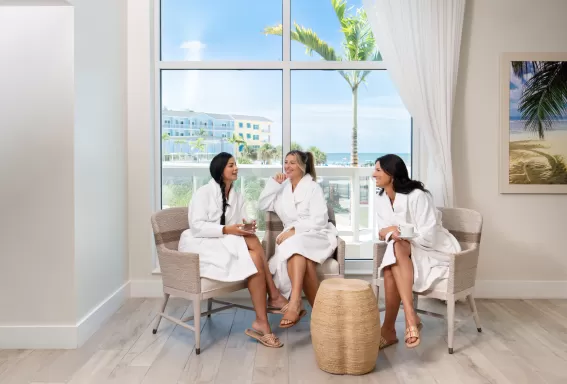 Three women wearing white spa robes sit in armchairs near a large window, talking and smiling while holding drinks, with palm trees, a beachfront resort building, and the ocean visible outside.